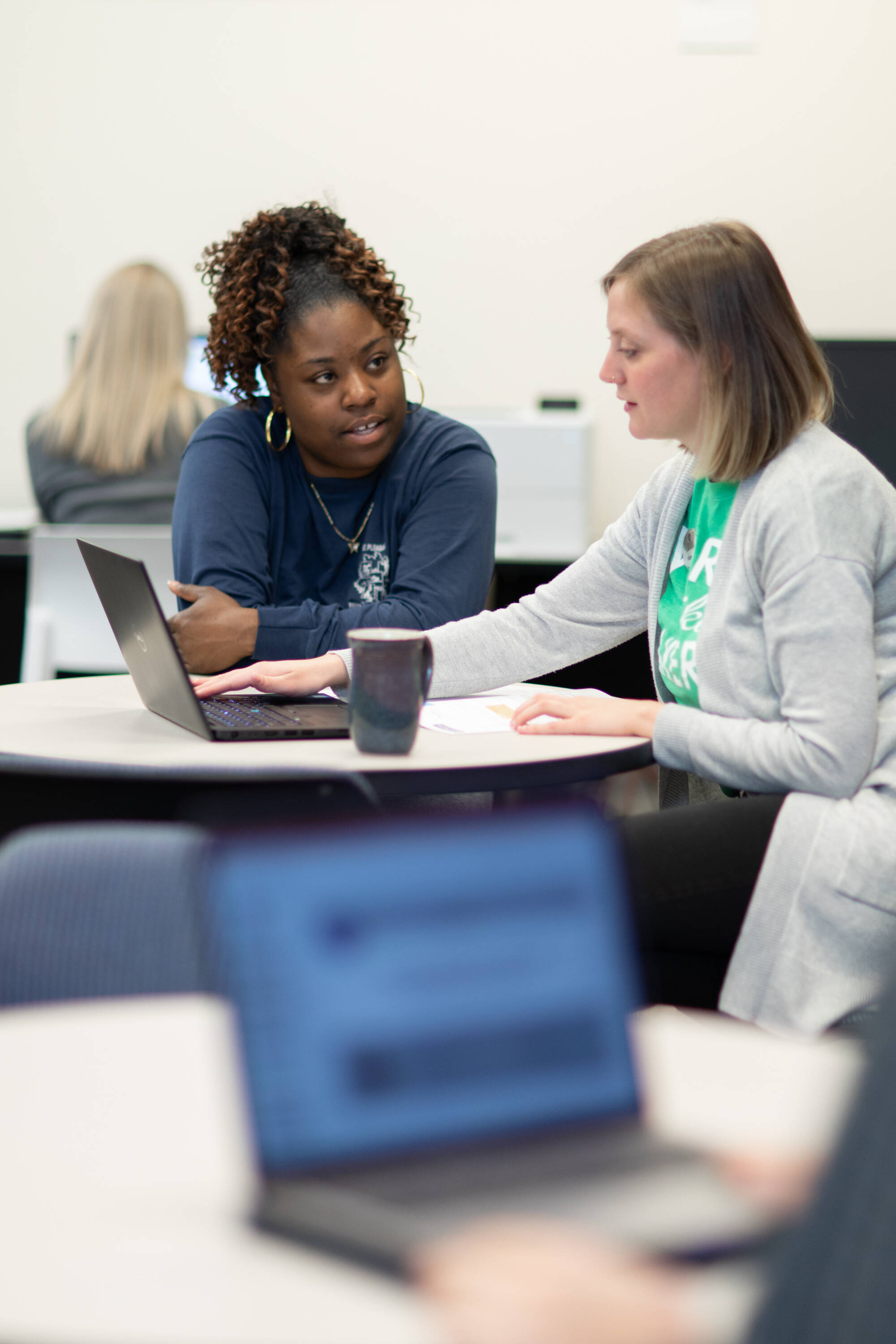 Two people collaborating at a table with a laptop in a classroom setting.